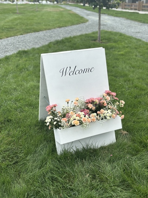 White A-board WELCOME sign with a floral bloom box on a grass lawn